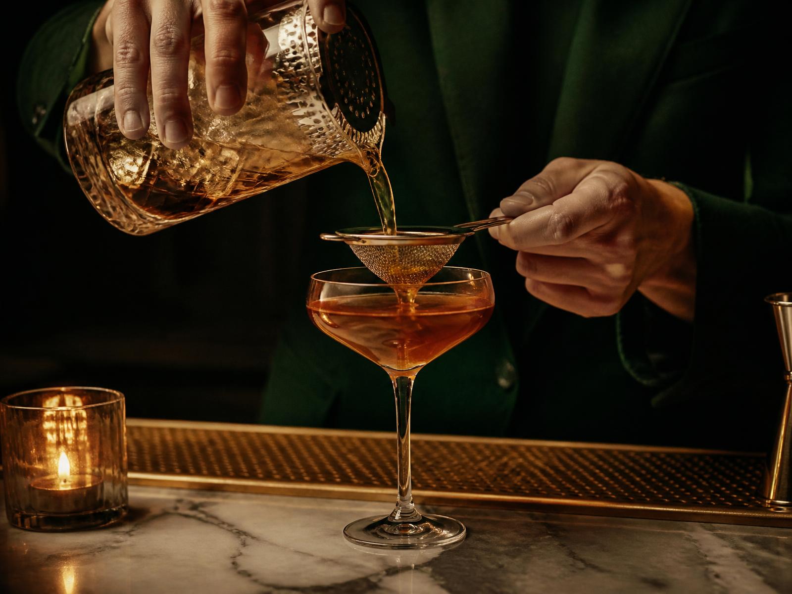 Bartender straining an amber cocktail into a coupe over a marble bar